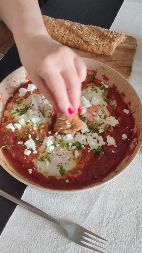 Finalno servirana shakshuka, rapsodija crvenih boja paprike i paradajiza u tiganju sa jajima, namrvljenim sirom i svezim korijandorom, uz to na drvenoj dasci serviran hleb